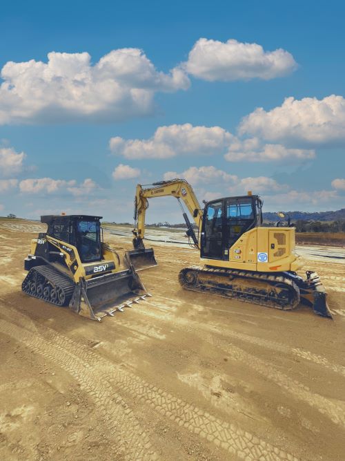 A bulldozer and an excavator parked on a dirt construction site under a blue sky with scattered clouds. The vehicles are on a sloped surface, with visible tracks in the dirt, indicating recent use.