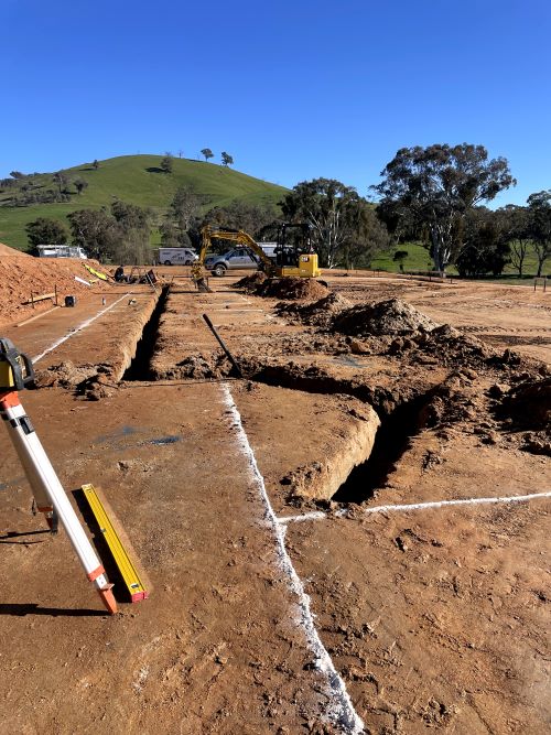 A construction site with trenches dug in a dirt field. White lines mark the ground, and a yellow excavator is in the background. Trees and a green hill are visible against a clear blue sky. A surveying tool is positioned in the foreground.