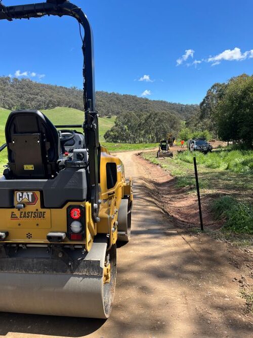 A yellow road roller sits on a dirt path surrounded by green grass and hilly landscape under a sunny blue sky. In the background, construction vehicles and a pickup truck are visible near the winding road.