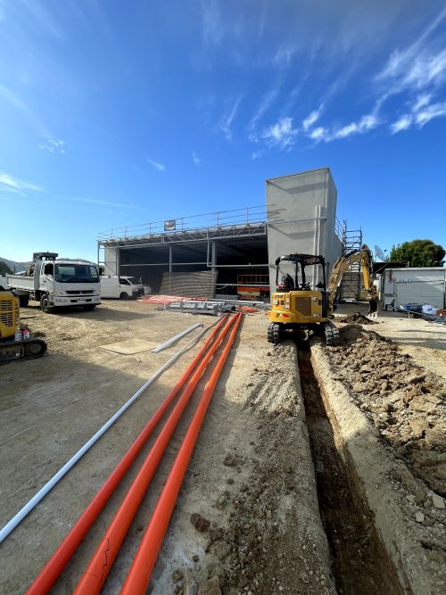 A construction site with a partially built structure in the background. In the foreground, there are trenches with orange pipes laid out. A small yellow excavator is parked nearby under a clear blue sky.