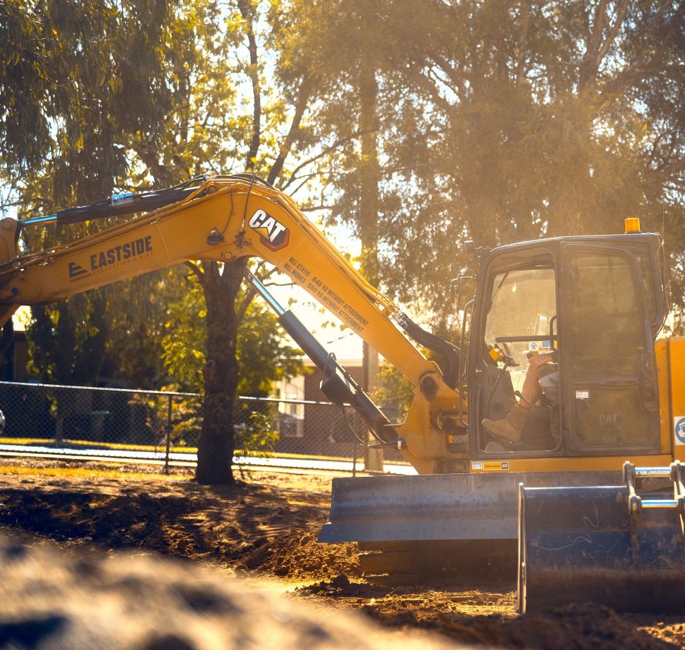 A construction worker operates a yellow excavator at a construction site. Sunlight filters through nearby trees, casting shadows. The machine is branded with "CAT" and "Eastside." A fence surrounds the area in the background.
