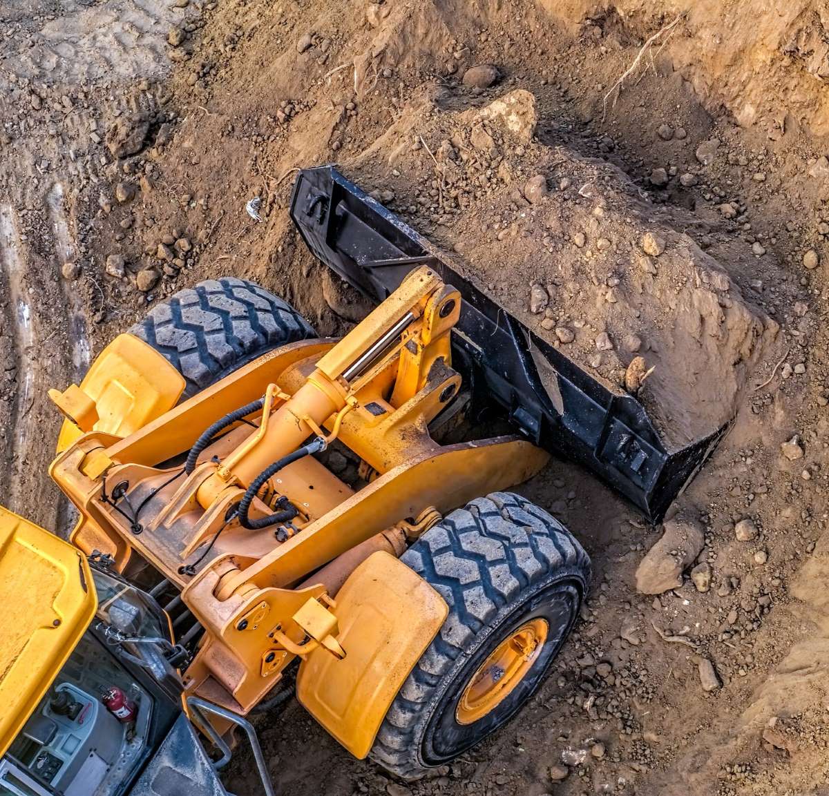 A yellow bulldozer with large wheels is moving a pile of dirt. The image is taken from above, showing the vehicle's bucket loaded with soil. The surrounding area is covered with loose dirt and earth.