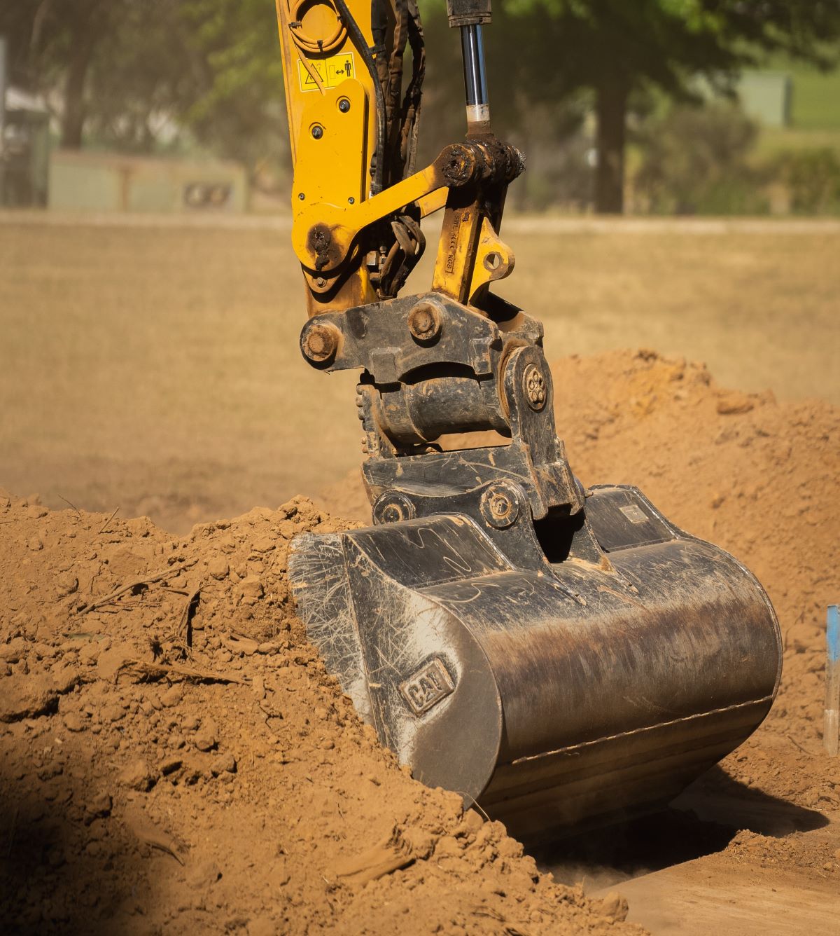 A close-up view of an excavator's yellow arm with a large metal bucket digging into a mound of dirt at a construction site, with blurred trees and grass in the background.
