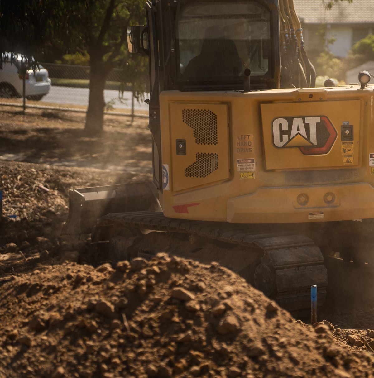 A construction vehicle branded "CAT" operates on a dusty construction site, surrounded by dirt and debris. Trees and a parked car are visible in the background.