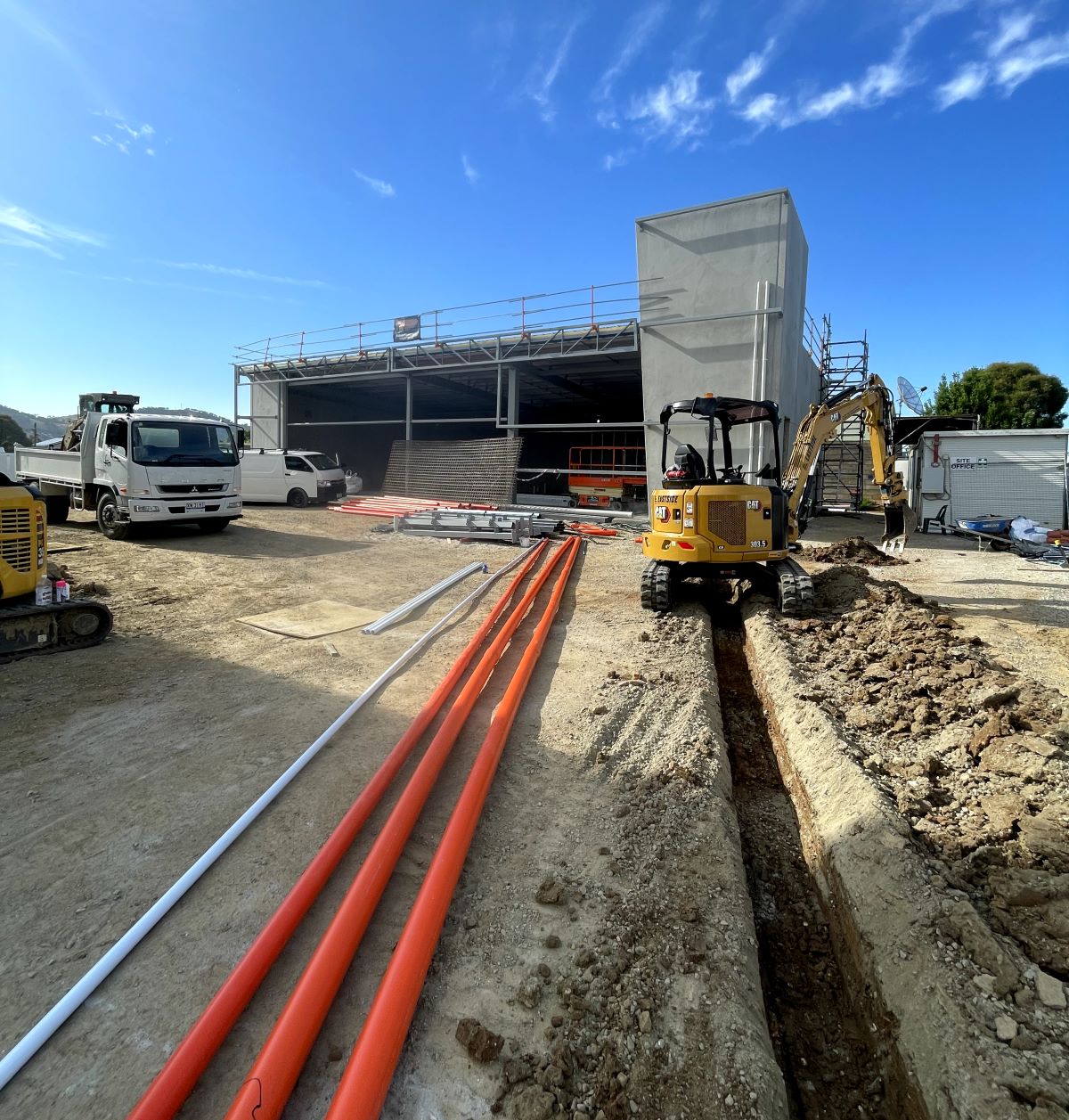 Construction site with a partially built structure, several vehicles, and an excavator digging a trench. Orange pipes are laid out on the ground under a clear blue sky.
