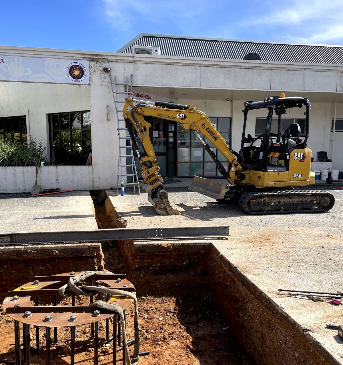 A small CAT excavator is parked near a building, next to a trench under construction. A ladder leans against the building, and construction tools and materials are scattered around the site.