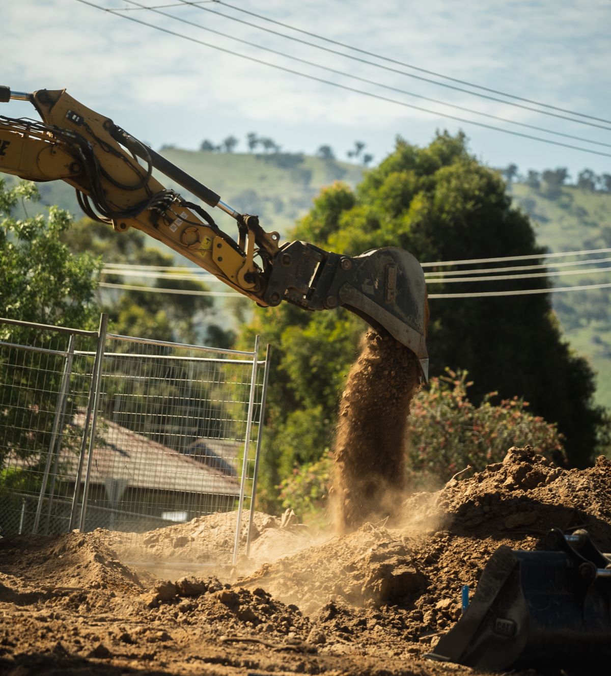A yellow excavator lifts a bucket of dirt at a construction site. Dust clouds form as the soil is dumped onto a mound. Trees and a hill with power lines are visible in the background under a partly cloudy sky.