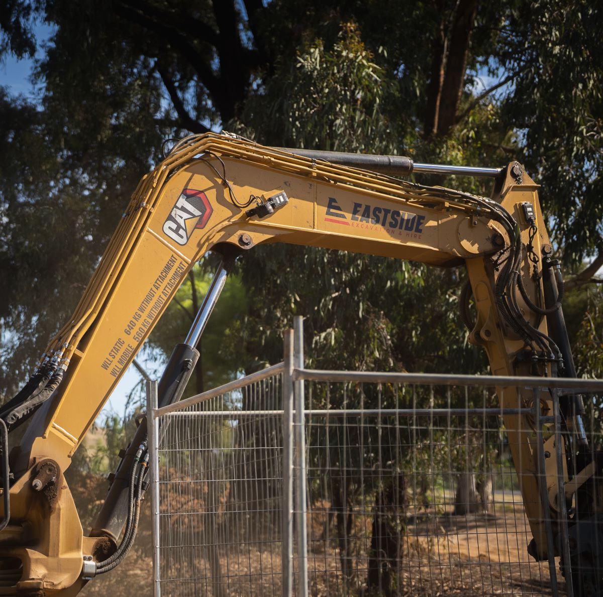 A construction excavator with a visible arm and hydraulic mechanism is parked behind a metal fence. There are trees in the background under a clear sky.