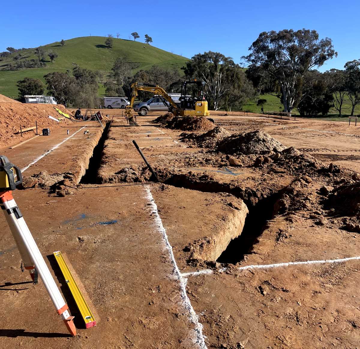 A construction site with dug trenches and marked outlines on the soil. An excavator is in the background, surrounded by green hills and trees under a clear blue sky. Construction tools are visible in the foreground.